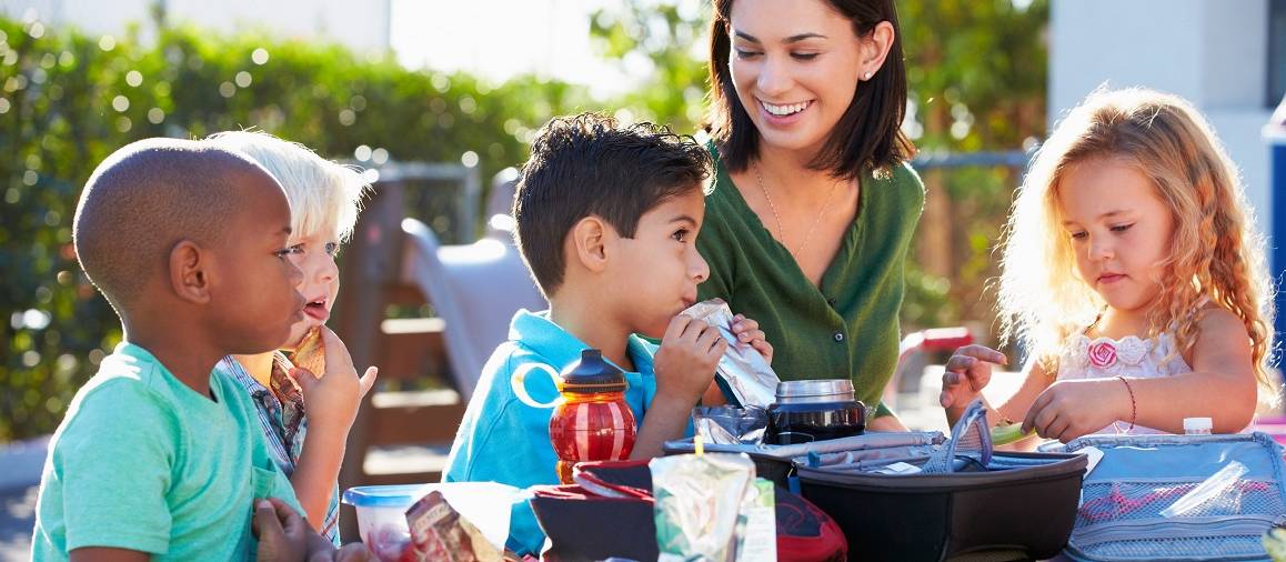 De vrijwilliger assisteert een pedagogisch medewerker van de school tijdens het lunchmoment met ongeveer 25 kinderen